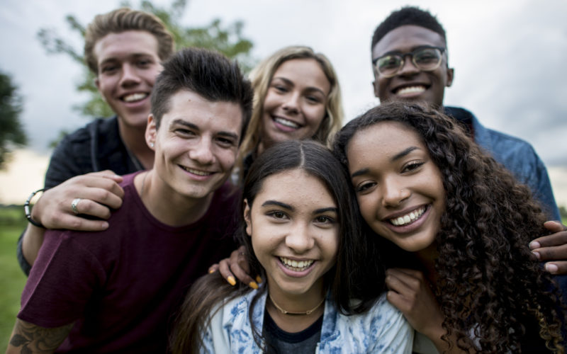 A multi-ethnic group of teenagers are outdoors on a cloudy day. They are wearing casual clothing. They are smiling while taking a selfie together.